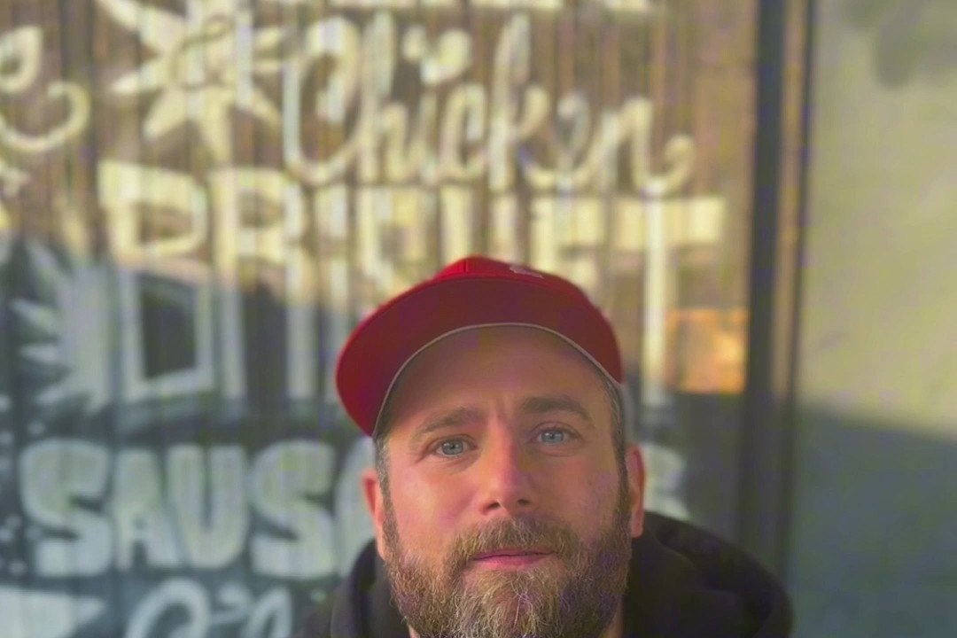 Philipp vor der Holzhütte, an der Wand die Kreideschrift Chicken, Brisket, Sausages